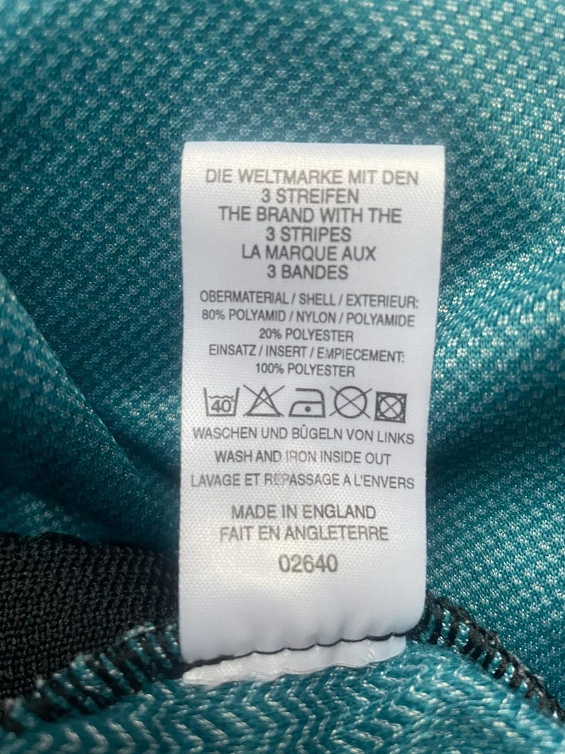 May include: White tag with black text and a washing instruction symbol. The text reads "Die Weltmarke mit den 3 Streifen The Brand with the 3 Stripes La Marque aux 3 Bandes Obermaterial/Shell/Exterieur: 80% Polyamid/Nylon/Polyamide 20% Polyester Einsatz/Insert/Empiecement: 100% Polyester Waschen und Bugeln von Links Wash and Iron Inside Out Lavage et Repassage a l'Envers Made in England Fait en Angleterre 02640"