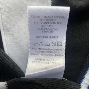 May include: A white tag with black text, featuring the phrases "DIE WELTMARKE MIT DEN 3 STREIFEN", "THE BRAND WITH THE 3 STRIPES", and "LA MARQUE AUX 3 BANDES". The tag also states "100% POLYESTER" and "MADE IN ENGLAND".