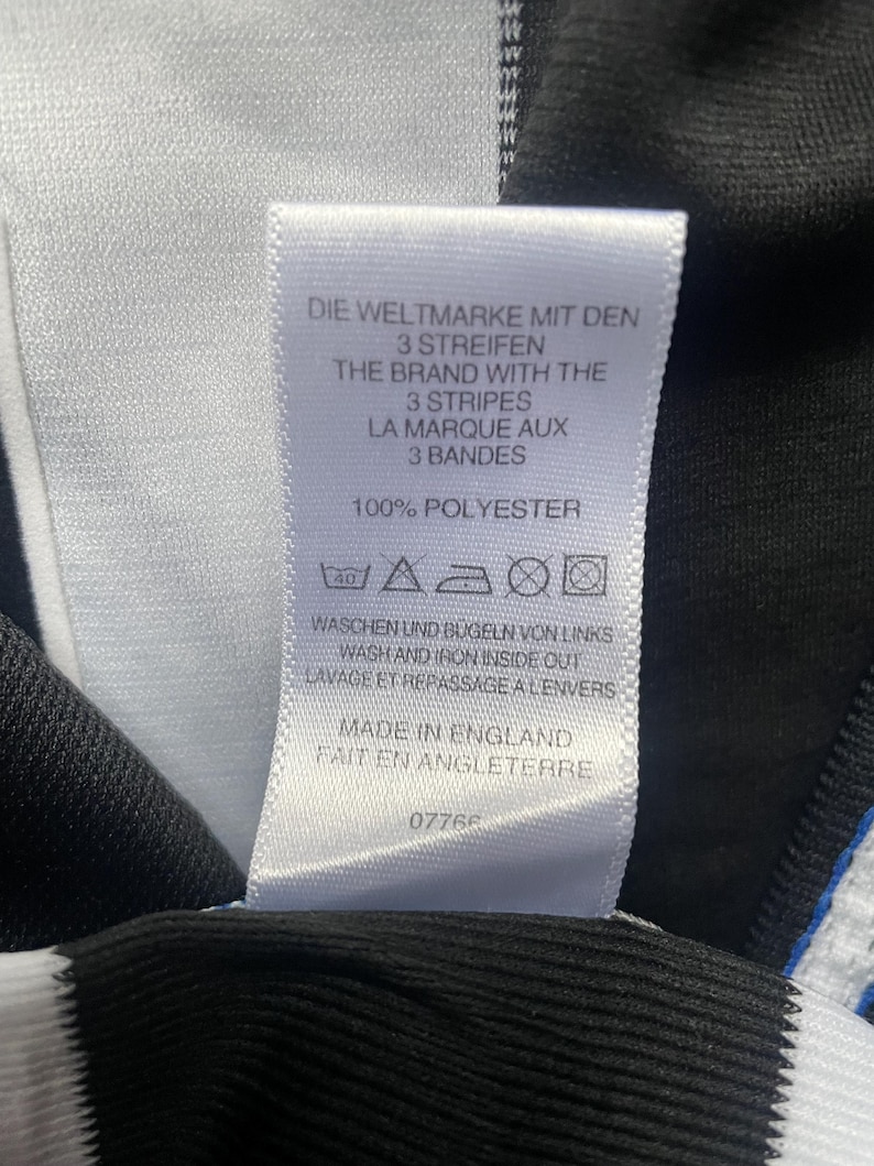 May include: A white tag with black text, featuring the phrases "DIE WELTMARKE MIT DEN 3 STREIFEN", "THE BRAND WITH THE 3 STRIPES", and "LA MARQUE AUX 3 BANDES". The tag also states "100% POLYESTER" and "MADE IN ENGLAND".