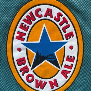 K&ouml;nnte beinhalten: Ein tealfarbenes Trikot mit einem gro&szlig;en, ovalen Aufn&auml;her. Der Aufn&auml;her zeigt die Worte "NEWCASTLE BROWN ALE" in roter Schrift, einen goldenen Rand, einen blauen Stern und eine Stadtsilhouette.