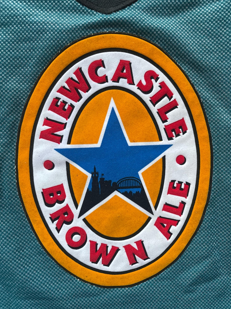K&ouml;nnte beinhalten: Ein tealfarbenes Trikot mit einem gro&szlig;en, ovalen Aufn&auml;her. Der Aufn&auml;her zeigt die Worte "NEWCASTLE BROWN ALE" in roter Schrift, einen goldenen Rand, einen blauen Stern und eine Stadtsilhouette.