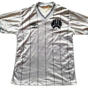 May include: A white and black striped soccer jersey with a black and white logo on the front. The logo says "MUF" and has a bull's head inside. The jersey has a v-neck and is made by Umbro.
