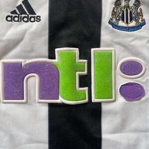 May include: A white and black striped football shirt with the Adidas logo, the Newcastle United crest, and the NTL logo in purple, green, and white.