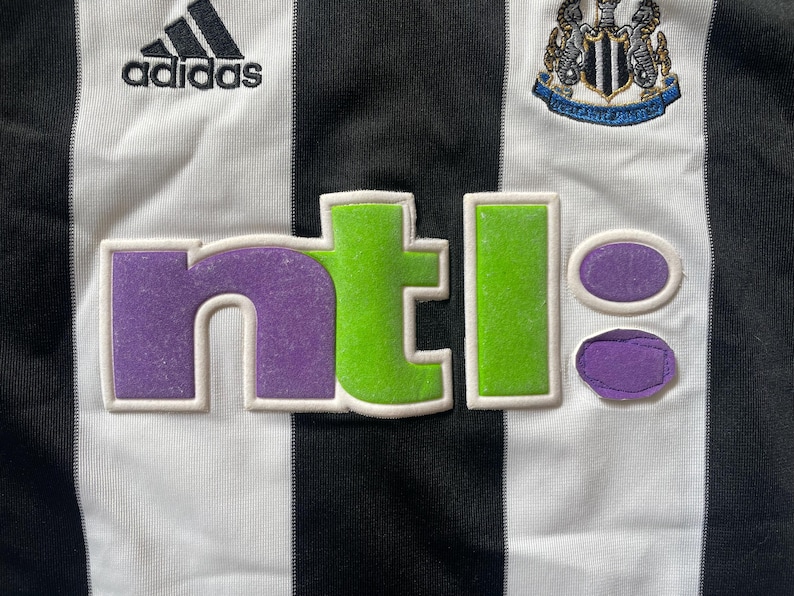 May include: A white and black striped football shirt with the Adidas logo, the Newcastle United crest, and the NTL logo in purple, green, and white.