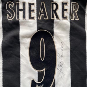 Puede incluir: Camiseta de f&uacute;tbol a rayas blancas y negras con el nombre "Shearer" y el n&uacute;mero "9" en la espalda. La camiseta tiene el logotipo de la FA Premier League. La camiseta est&aacute; firmada por Alan Shearer.