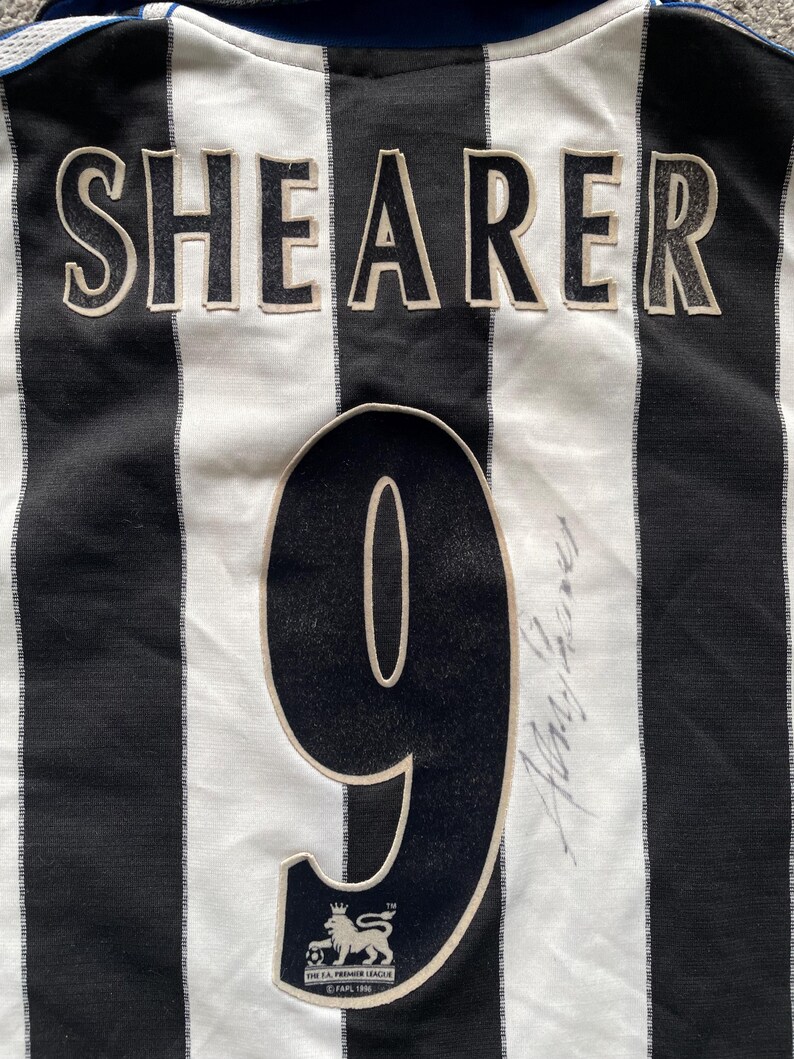 Puede incluir: Camiseta de f&uacute;tbol a rayas blancas y negras con el nombre "Shearer" y el n&uacute;mero "9" en la espalda. La camiseta tiene el logotipo de la FA Premier League. La camiseta est&aacute; firmada por Alan Shearer.