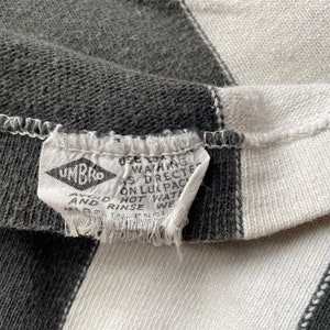 Puede incluir: Primer plano de una tela a rayas blancas y negras con una etiqueta blanca que dice "UMBRO" y "USE COLD WASHING AS DIRECTED ON LUX PACK HOT WATER AND RINSE WELL."