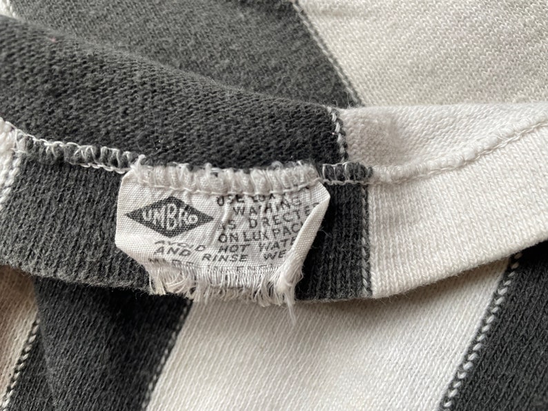 Puede incluir: Primer plano de una tela a rayas blancas y negras con una etiqueta blanca que dice "UMBRO" y "USE COLD WASHING AS DIRECTED ON LUX PACK HOT WATER AND RINSE WELL."
