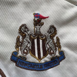 May include: Close-up of a white fabric with the embroidered Newcastle United crest. The crest includes seahorses, a shield with stripes, and a blue banner with the team's name.