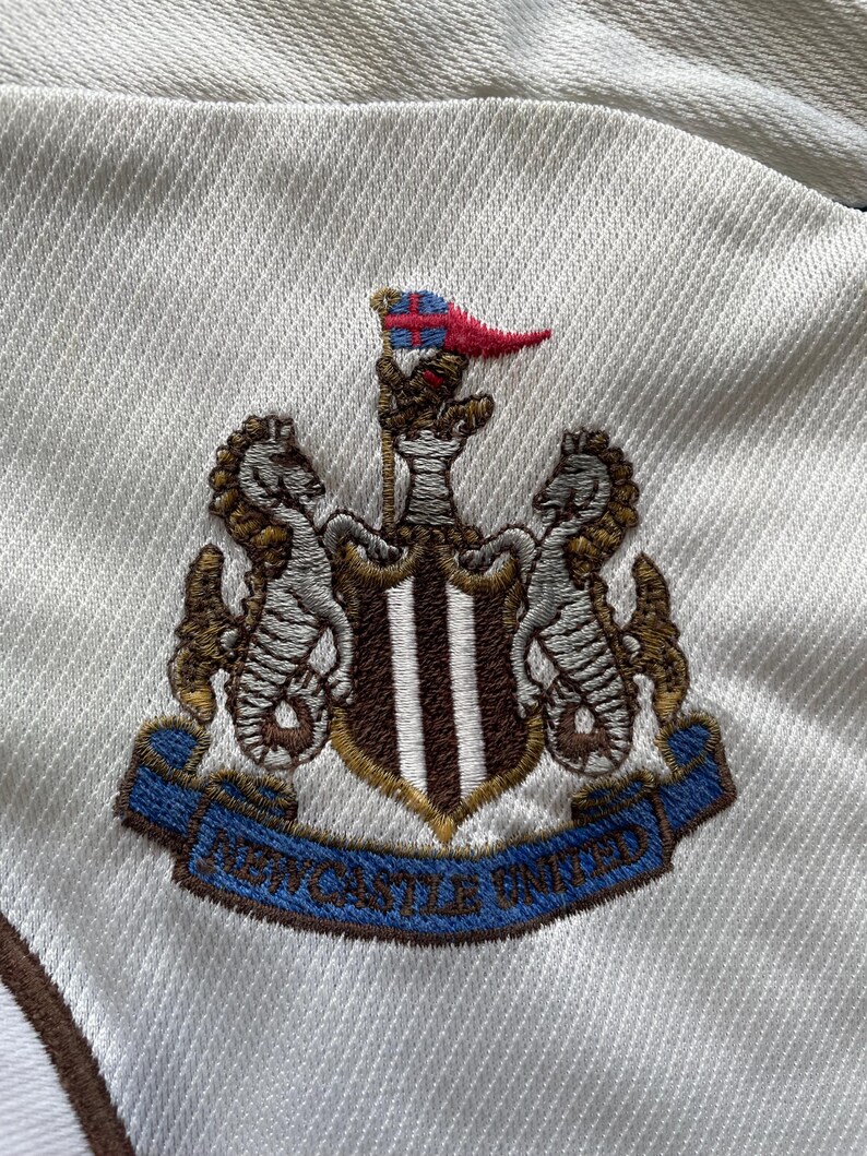May include: Close-up of a white fabric with the embroidered Newcastle United crest. The crest includes seahorses, a shield with stripes, and a blue banner with the team's name.