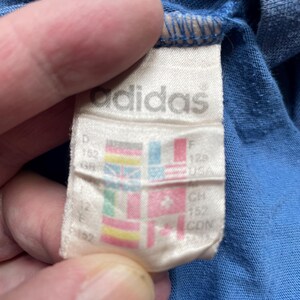 May include: White tag with the word "adidas" printed in black. The tag has a size chart with different country sizes, including D 152, F 128, CH 152, CDN 152.