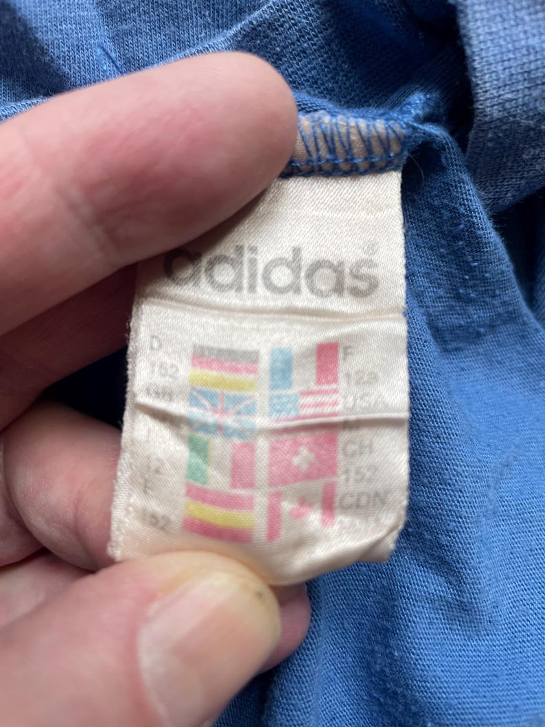 May include: White tag with the word "adidas" printed in black. The tag has a size chart with different country sizes, including D 152, F 128, CH 152, CDN 152.