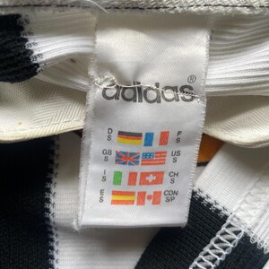 May include: Close-up of a white Adidas tag with the Adidas logo and various country flags. The tag is attached to a black and white striped fabric. The tag includes size information for different regions.