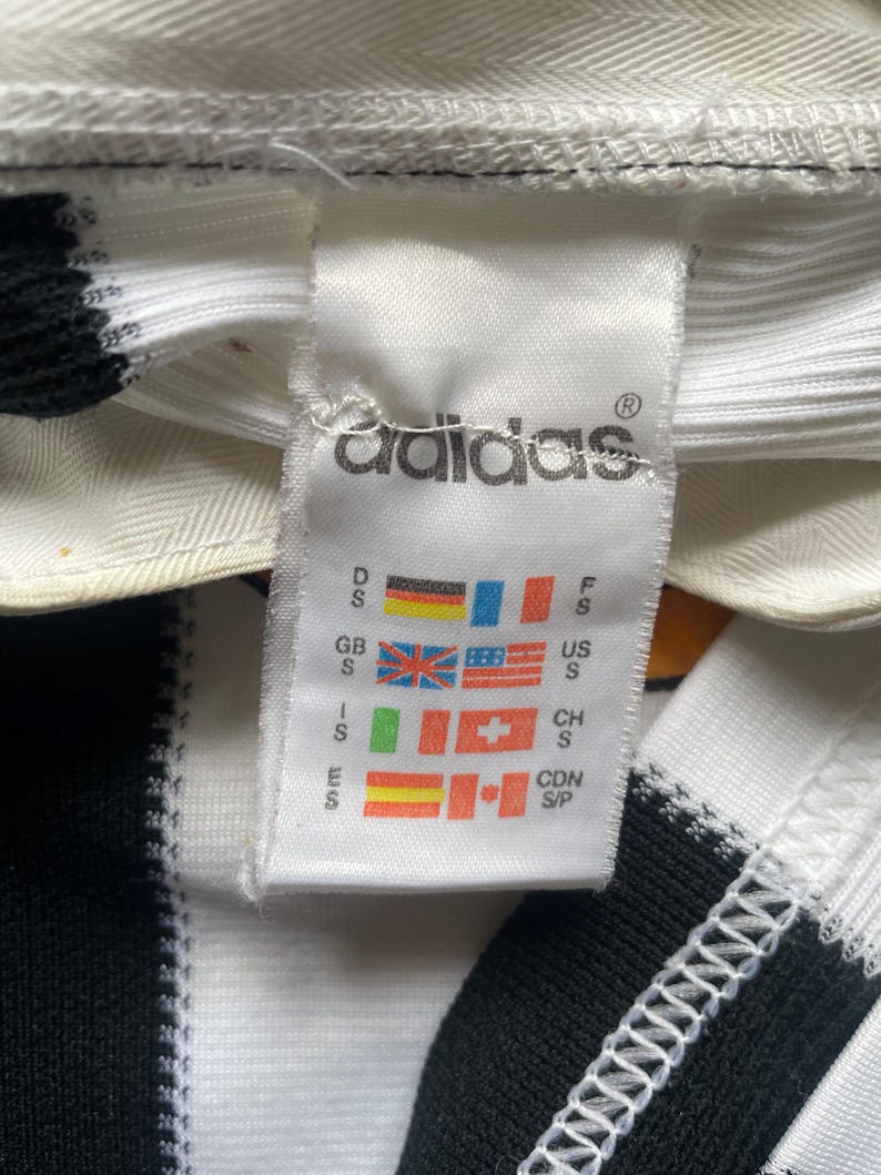 May include: Close-up of a white Adidas tag with the Adidas logo and various country flags. The tag is attached to a black and white striped fabric. The tag includes size information for different regions.