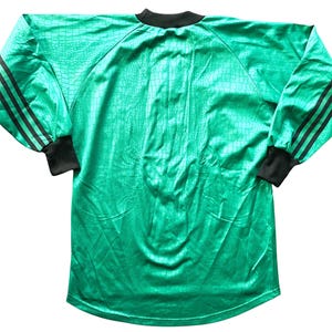 May include: A long-sleeved, emerald green athletic jersey with a textured pattern. The jersey features black stripes on the sleeves and black cuffs and collar. The jersey is made of a shiny material.