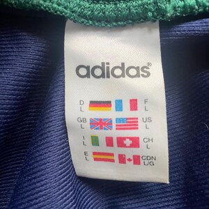 May include: A white Adidas tag with the brand name and size indicators. The tag features flags representing different countries, including the United States, Great Britain, and Canada, along with size designations.