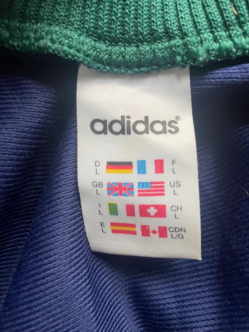 May include: A white Adidas tag with the brand name and size indicators. The tag features flags representing different countries, including the United States, Great Britain, and Canada, along with size designations.
