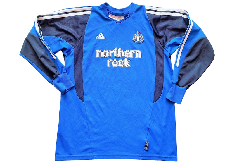 May include: A blue long-sleeved soccer jersey with black and white accents. The jersey has the Adidas logo, the Newcastle United crest, and the text "northern rock" on the front.