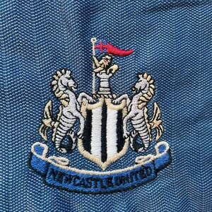 May include: A black and white embroidered crest with a shield, two seahorses, and a banner that reads "NEWCASTLE UNITED".