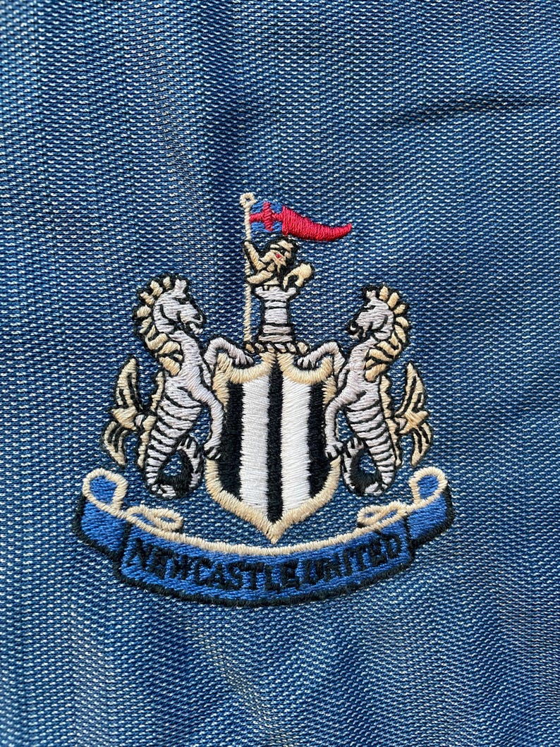 May include: A black and white embroidered crest with a shield, two seahorses, and a banner that reads "NEWCASTLE UNITED".