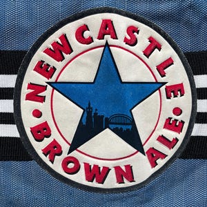 May include: A circular patch featuring the words "Newcastle Brown Ale" in red, with a blue star and city silhouette. The patch is set against a blue fabric background with black and white stripes.