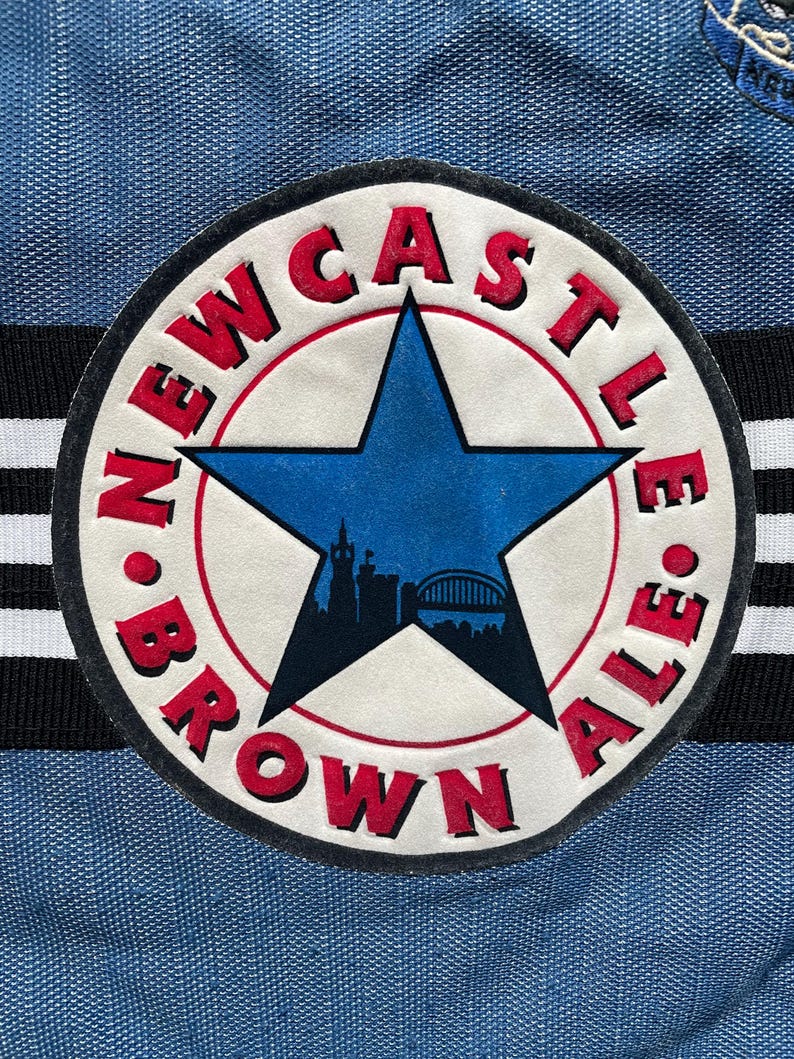 May include: A circular patch featuring the words "Newcastle Brown Ale" in red, with a blue star and city silhouette. The patch is set against a blue fabric background with black and white stripes.