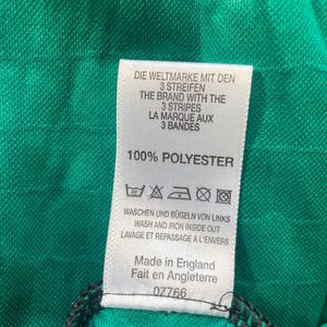 May include: Close-up of a green fabric with a tag. The tag reads "DIE WELTMARKE MIT DEN 3 STREIFEN" and "THE BRAND WITH THE 3 STRIPES". It also says "100% POLYESTER" and "Made in England".