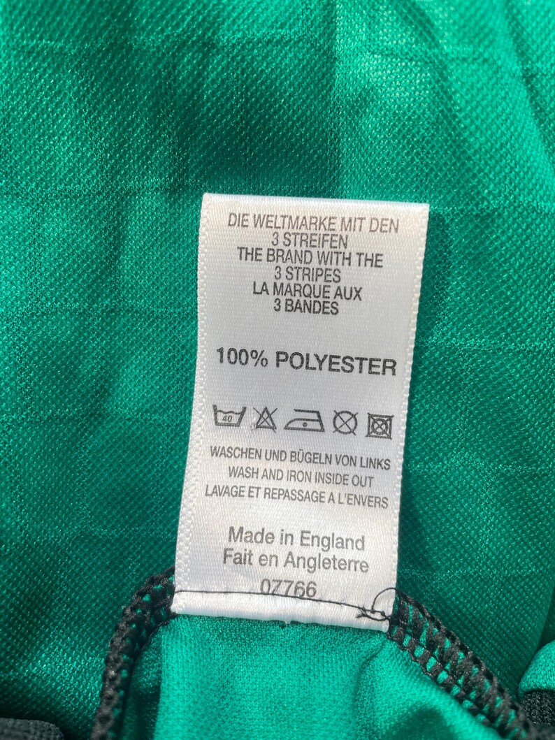 May include: Close-up of a green fabric with a tag. The tag reads "DIE WELTMARKE MIT DEN 3 STREIFEN" and "THE BRAND WITH THE 3 STRIPES". It also says "100% POLYESTER" and "Made in England".