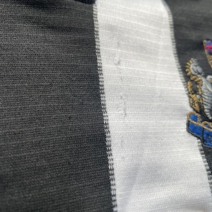 May include: Black and white striped football shirt with a blue collar and a team crest on the right side. The crest features a bird with a shield and a crown.