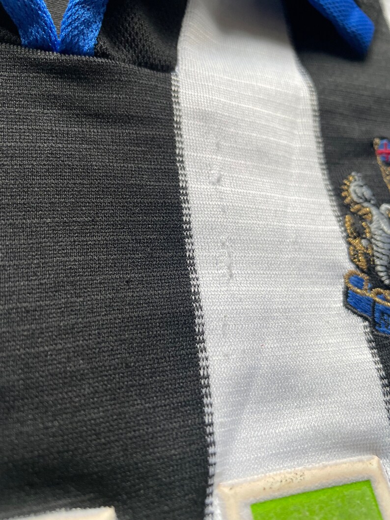 May include: Black and white striped football shirt with a blue collar and a team crest on the right side. The crest features a bird with a shield and a crown.