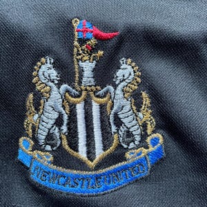 May include: Embroidered black and white crest with a gold outline and a blue banner reading "NEWCASTLE UNITED".