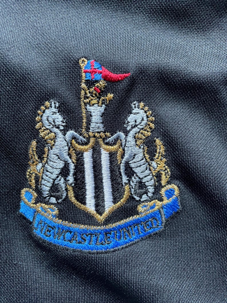 May include: Embroidered black and white crest with a gold outline and a blue banner reading "NEWCASTLE UNITED".