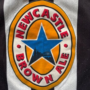 May include: Close-up of a black and white striped jersey featuring a Newcastle Brown Ale logo. The logo is a circular design with the words "NEWCASTLE BROWN ALE" in red lettering around a blue star and a city silhouette.