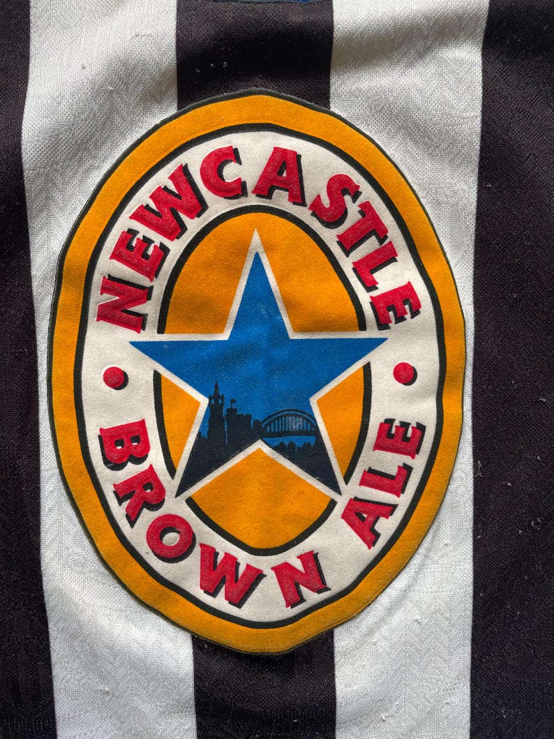May include: Close-up of a black and white striped jersey featuring a Newcastle Brown Ale logo. The logo is a circular design with the words "NEWCASTLE BROWN ALE" in red lettering around a blue star and a city silhouette.