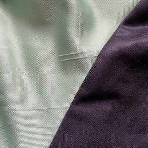 May include: Close-up of a section of fabric with a diagonal seam. The fabric is divided into two sections: a light green textured material and a dark purple, smooth material. The seam is stitched with white thread.
