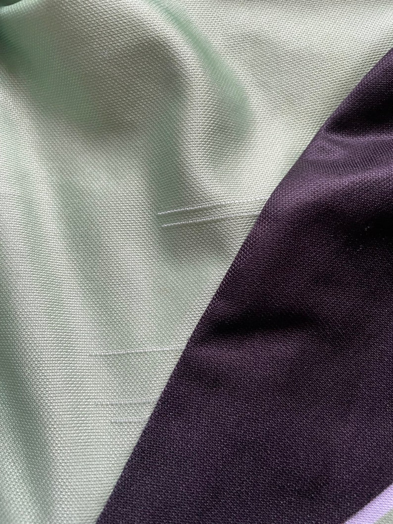 May include: Close-up of a section of fabric with a diagonal seam. The fabric is divided into two sections: a light green textured material and a dark purple, smooth material. The seam is stitched with white thread.