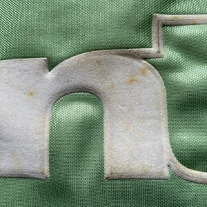 May include: Close-up of a green fabric with a white felt letter "n" sewn on it. The letter has a slightly worn appearance.