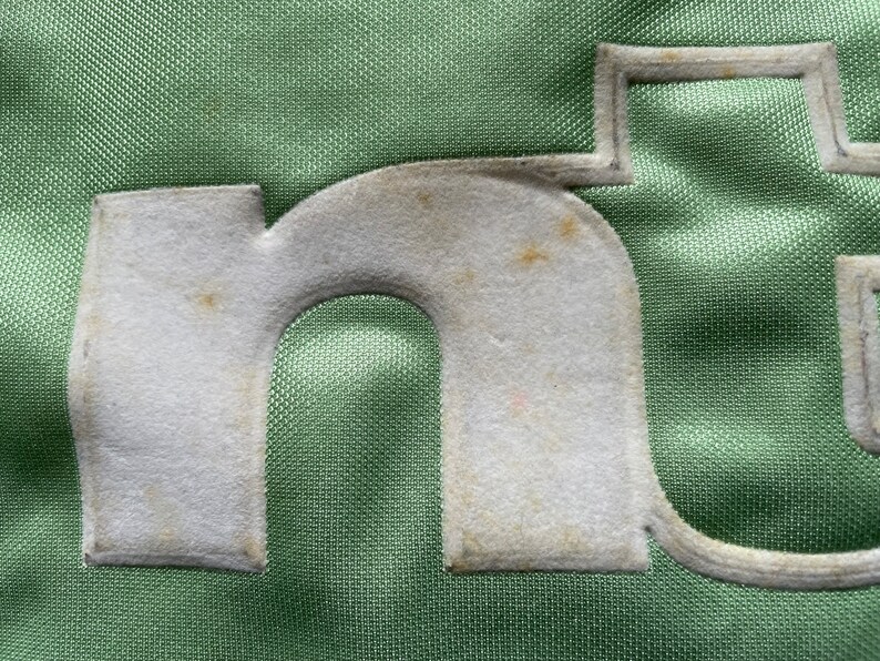May include: Close-up of a green fabric with a white felt letter "n" sewn on it. The letter has a slightly worn appearance.