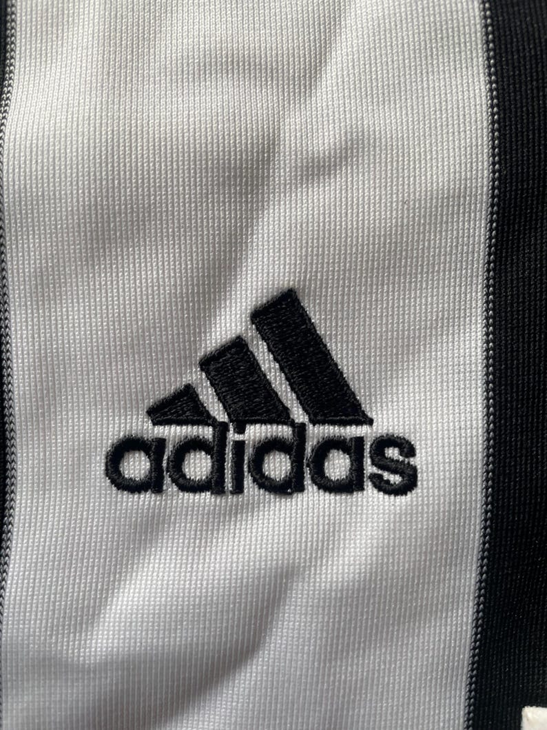 May include: A white and black striped athletic jersey with the Adidas logo and the word "adidas" in black.