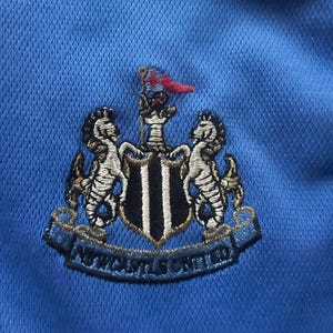 K&ouml;nnte beinhalten: Nahaufnahme eines hellblauen Stoffes mit einem gestickten Newcastle United-Wappen. Das Wappen zeigt zwei Seepferdchen, einen Schild mit schwarz-wei&szlig;en Streifen und den Text "NEWCASTLE UNITED".