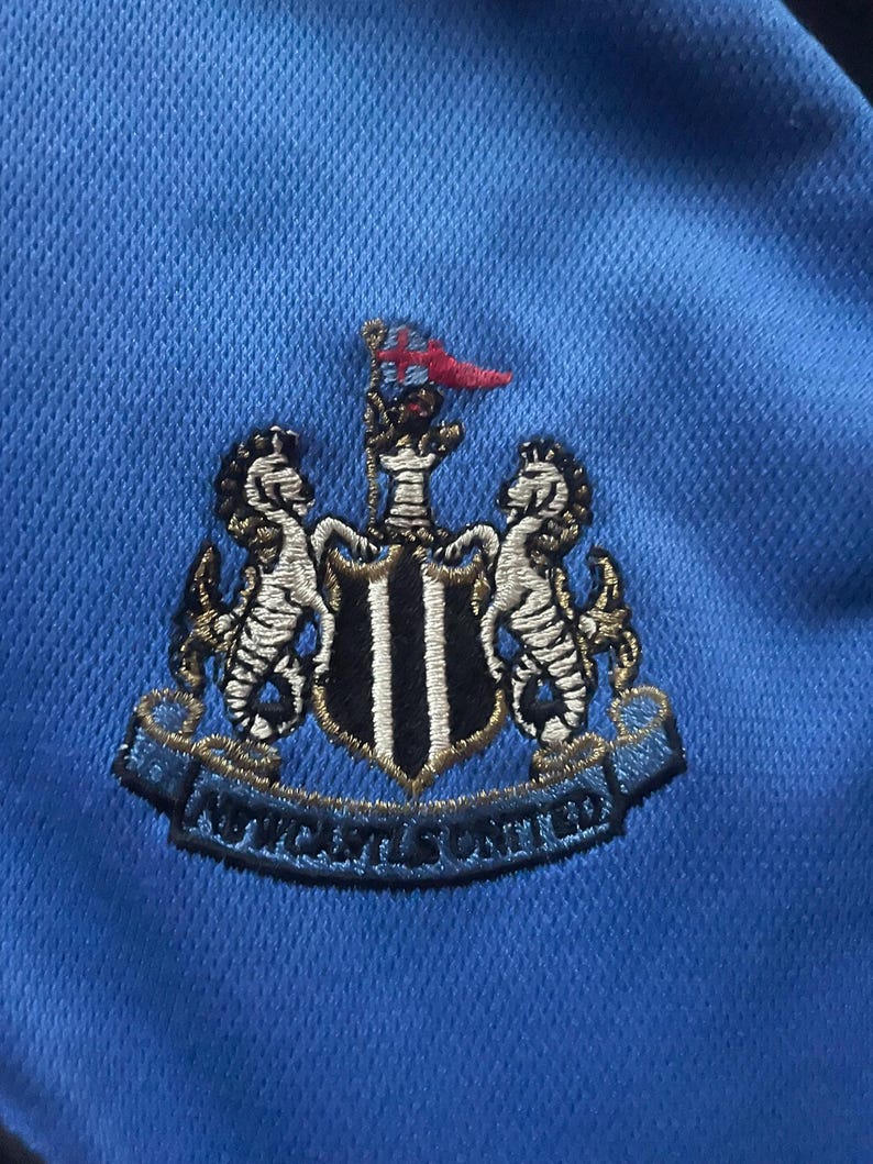 K&ouml;nnte beinhalten: Nahaufnahme eines hellblauen Stoffes mit einem gestickten Newcastle United-Wappen. Das Wappen zeigt zwei Seepferdchen, einen Schild mit schwarz-wei&szlig;en Streifen und den Text "NEWCASTLE UNITED".