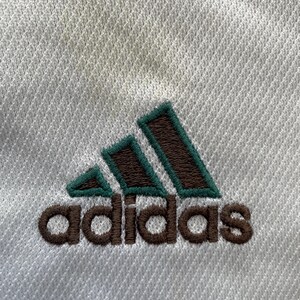 May include: Close-up of white athletic fabric with the Adidas logo embroidered in brown and green. The logo features three green angled stripes above the brand name.