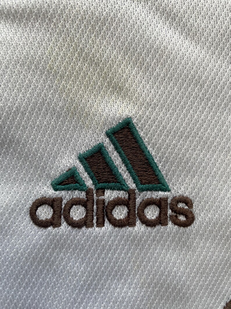 May include: Close-up of white athletic fabric with the Adidas logo embroidered in brown and green. The logo features three green angled stripes above the brand name.