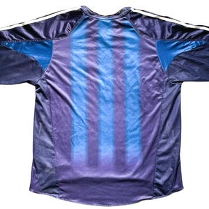 May include: A navy blue athletic jersey with blue and purple vertical stripes on the back. The jersey has a crew neck and short sleeves. White stripes run down the shoulders.
