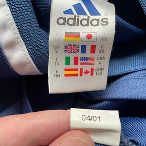 Op de afbeelding: Een blauw Adidas-shirt met een witte streep aan de zijkant en een maatlabel met het Adidas-logo en maatgegevens. Het label bevat ook een datum van 04/01.