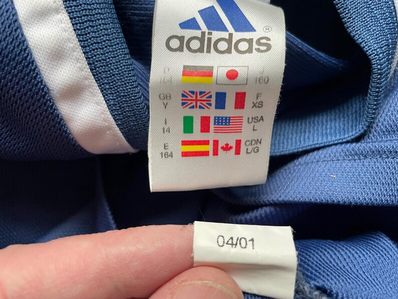 Op de afbeelding: Een blauw Adidas-shirt met een witte streep aan de zijkant en een maatlabel met het Adidas-logo en maatgegevens. Het label bevat ook een datum van 04/01.
