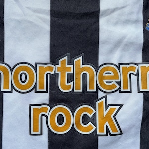 May include: A black and white striped soccer jersey with the words "northern rock" in yellow letters on the front. The jersey also features a team logo on the right side and the Adidas logo on the left side.