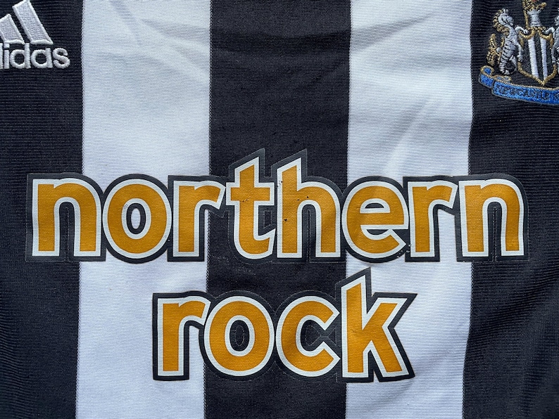 May include: A black and white striped soccer jersey with the words "northern rock" in yellow letters on the front. The jersey also features a team logo on the right side and the Adidas logo on the left side.