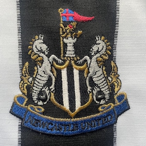 May include: Embroidered patch featuring the Newcastle United crest. The crest includes two seahorses, a shield with black and white stripes, and a banner with the words "Newcastle United". The background is a black and white striped pattern.