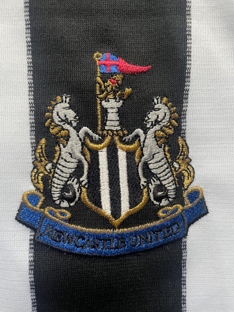 May include: Embroidered patch featuring the Newcastle United crest. The crest includes two seahorses, a shield with black and white stripes, and a banner with the words "Newcastle United". The background is a black and white striped pattern.